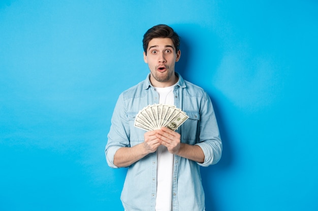 Hombre sorprendido sosteniendo dinero, mirando al frente se preguntó, de pie sobre la pared azul