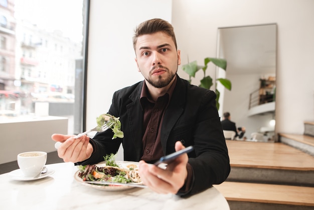 Un hombre sorprendido en un traje se sienta en un acogedor restaurante con un teléfono inteligente en sus manos, come una ensalada con un plato con un tenedor y mira a la cámara.