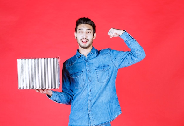 Hombre sosteniendo una caja de regalo plateada en la pared roja y mostrando un signo de mano positivo.