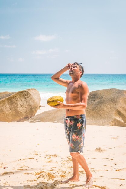 Foto hombre sosteniendo un coco mientras mira hacia la playa