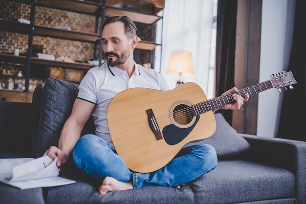 Hombre sosteniendo la guitarra y mirando notas mientras está sentado en el sofá en casa