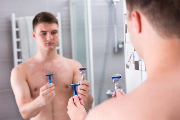 Hombre sosteniendo navajas de afeitar y elige lo mejor mientras está de pie frente al espejo en el moderno baño de azulejos en casa