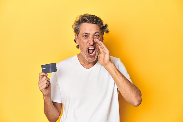 Hombre sosteniendo una tarjeta de crédito fondo de estudio amarillo gritando y sosteniendo la palma cerca de la boca abierta