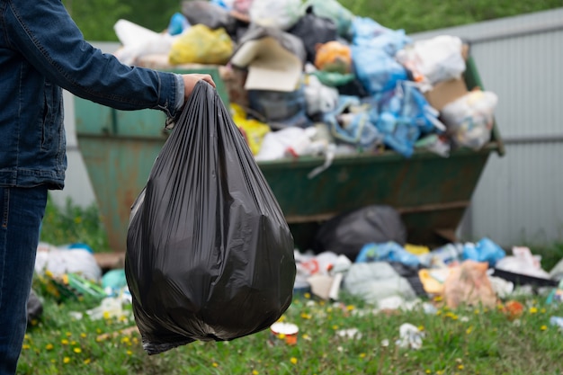 El hombre sostiene una bolsa de plástico llena de basura delante del contenedor de basura desbordante al aire libre en el campo