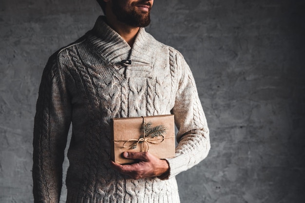 Un hombre sostiene una caja de artesanía para un regalo de Navidad con un suéter beige sobre un fondo gris