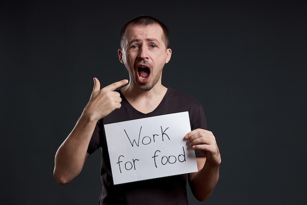 El hombre sostiene una hoja de papel de cartel en sus manos con la inscripción Trabajo por la comida. Sonrisa y alegría, lugar para texto, espacio de copia