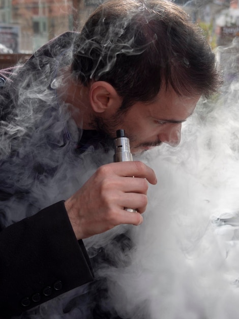 Un hombre sostiene en su mano y fuma un cigarrillo electrónico VAPE y libera nubes de humo de vapor