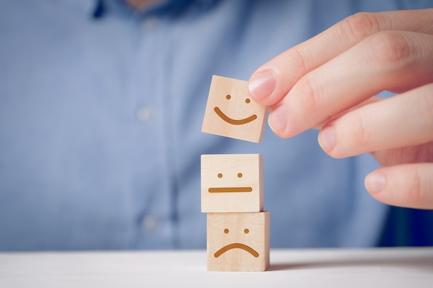 Un hombre sostiene con sus dedos un cubo de madera con una cara positiva al lado de uno descontento y neutral. para evaluar una acción o recurso.