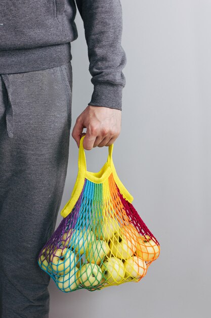 Un hombre sostiene en sus manos una bolsa de hilo de algodón reutilizable, un arco iris multicolor con manzanas verdes en su interior. fondo gris. Concepto de desperdicio cero