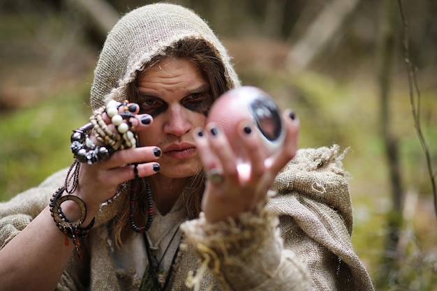 Un hombre con una sotana pasa un ritual en un bosque oscuro con una bola de cristal y un libro.