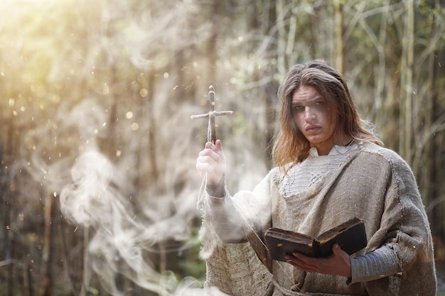 Un hombre con una sotana pasa un ritual en un bosque oscuro con una bola de cristal y un libro.