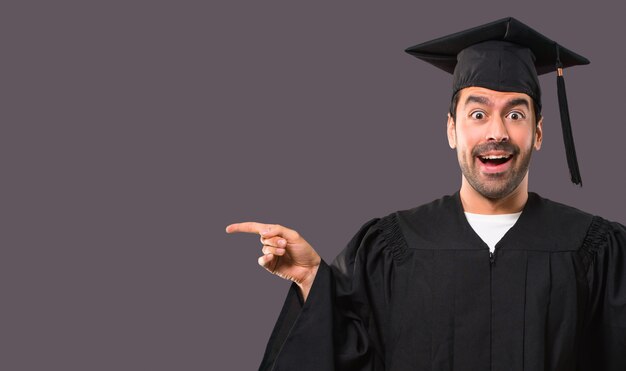 Hombre en su día de graduación Universidad apuntando el dedo a un lado y presentando un producto