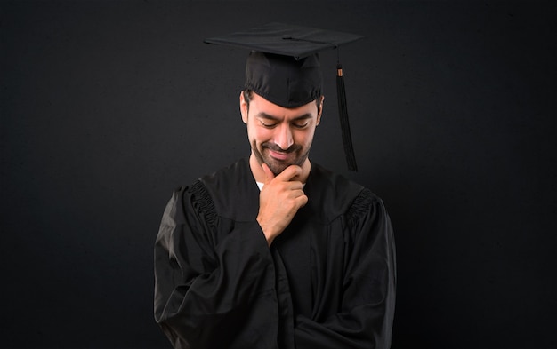 Hombre en su día de graduación Universidad de pie y mirando hacia abajo con la mano en el mentón