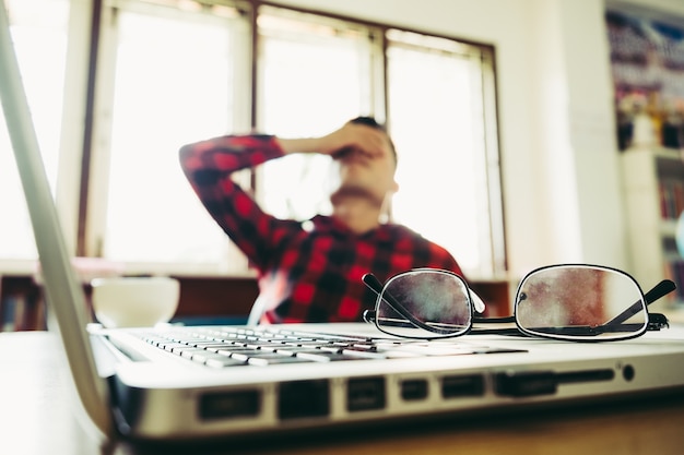 hombre con su mano sosteniendo su cara tomando un freno de trabajar con ordenador portátil y portátil con gafas en el escritorio de madera. concepto de estrés / descanso / tensión / fracaso / desaliento / depresión