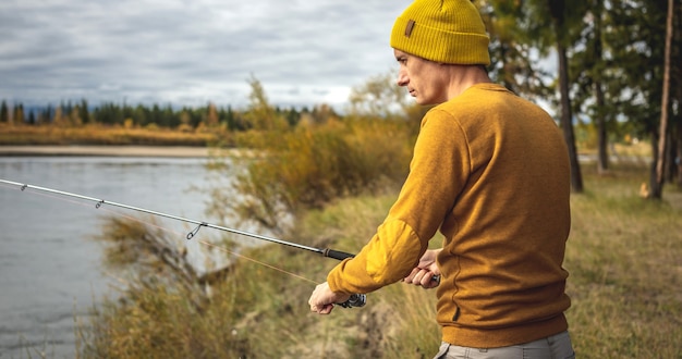 El hombre con un suéter amarillo y un sombrero está pescando en la orilla del río en el bosque de otoño con una varilla giratoria en sus manos