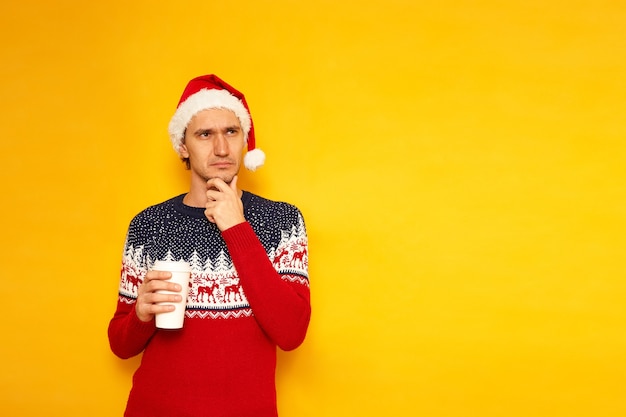 El hombre en el suéter rojo de Año Nuevo con gorro de Papá Noel con una taza de café de papel tiene su mano en la barbilla piensa