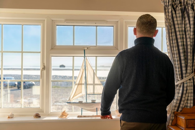 Hombre con suéter sentado cerca de la ventana en una clásica habitación de hotel antigua con vista al mar