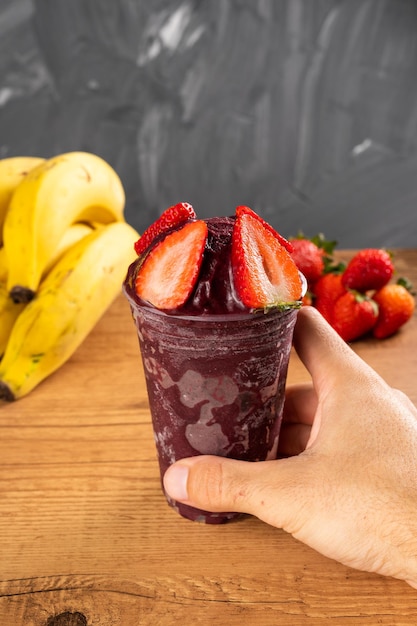 Hombre sujetando un batido helado brasileño AÃ§ai Berry helado en vaso de plástico con fresas. Sobre un escritorio de madera y un fondo de verano gris con frutas. Vista frontal para menú y redes sociales.