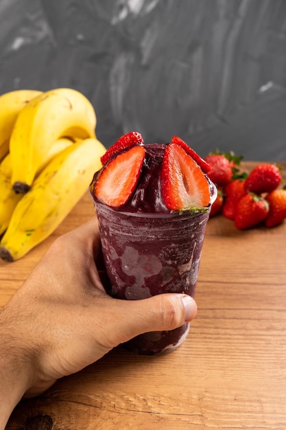 Hombre sujetando un batido helado brasileño AÃ§ai Berry helado en vaso de plástico con fresas. Sobre un escritorio de madera y un fondo de verano gris con frutas. Vista frontal para menú y redes sociales.