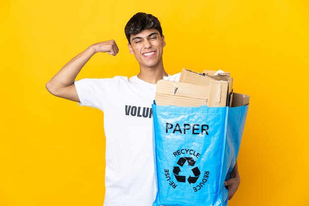 Hombre sujetando una bolsa de reciclaje llena de papel para reciclar sobre fondo amarillo aislado haciendo un gesto fuerte