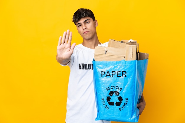 Hombre sujetando una bolsa de reciclaje llena de papel para reciclar sobre pared amarilla aislada haciendo gesto de parada