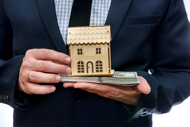 Hombre sujetando el modelo de casa de madera y billetes de dólar