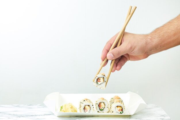 Hombre sujetando palillos y comiendo sushi japonés en plato desechable.