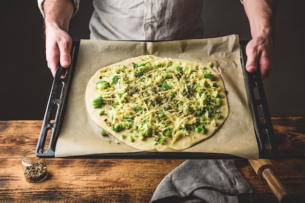 Hombre sujetando un plato para hornear con pizza cruda con brócoli, salsa pesto, especias y queso