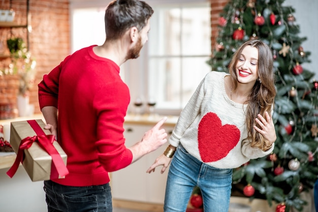 Hombre sujetando un regalo detrás para una mujer joven que celebra las vacaciones de Año Nuevo en casa