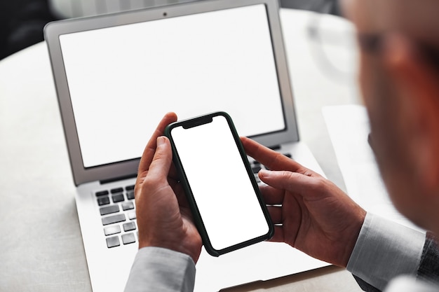 Hombre sujetando un teléfono móvil con una pantalla blanca en el fondo de una computadora portátil con una pantalla blanca