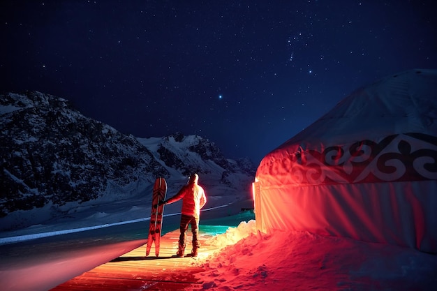 Un hombre con una tabla de snowboard parado cerca de una yurta por la noche en invierno en las montañas Almaty Kazajstán