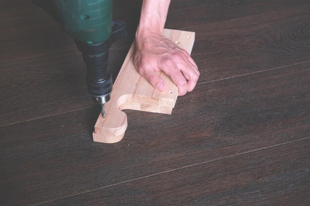 Un hombre taladra un agujero en un detalle de madera para hacer muebles hechos a manoFabricación de muebles en casa