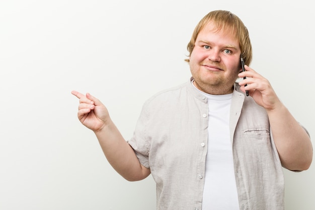 Hombre de talla grande de raza caucásica llamando por teléfono sonriendo alegremente apuntando con el dedo índice.