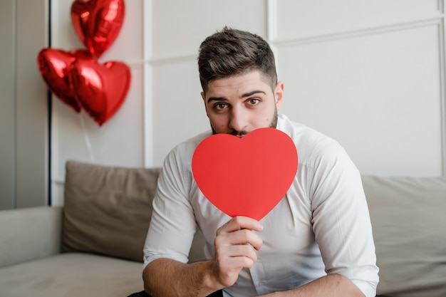 Foto hombre con tarjeta de san valentín en forma de corazón rojo sentado en el sofá con globos en casa