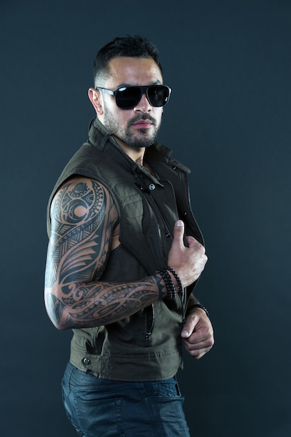 Hombre tatuado con bíceps y tríceps. Modelo de tatuaje con barba en la cara sin afeitar. Hombre barbudo con tatuaje en brazos fuertes. Macho de moda en gafas de sol de moda. Estilo de moda y tendencia.