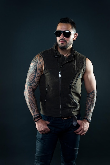 Hombre tatuado con bíceps y tríceps Modelo de tatuaje con barba en la cara sin afeitar Hombre barbudo con tatuaje en brazos fuertes Macho de moda con gafas de sol de moda Estilo de moda y tendencia