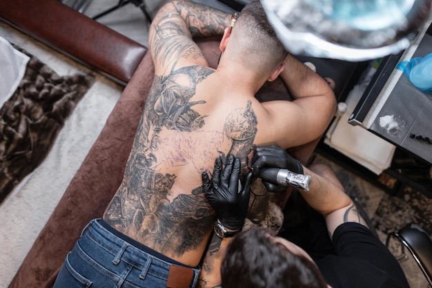 Hombre tatuador profesional con guantes negros y tatuaje de máquina de tatuaje en la espalda de un hombre, vista superior. Flujo de trabajo de creación de dibujos