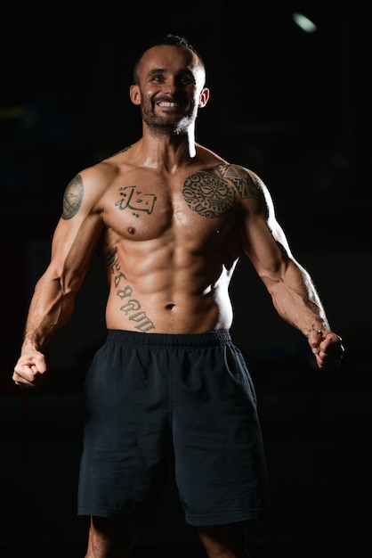 Hombre de tatuaje joven sano de pie fuerte en el gimnasio y flexionando los músculos Muscular culturista atlético modelo de fitness posando después de los ejercicios