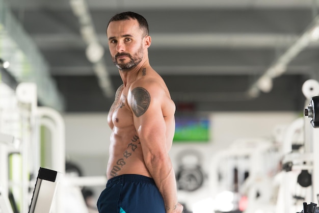 Foto hombre de tatuaje joven sano de pie fuerte en el gimnasio y flexionando los músculos muscular culturista atlético modelo de fitness posando después de los ejercicios