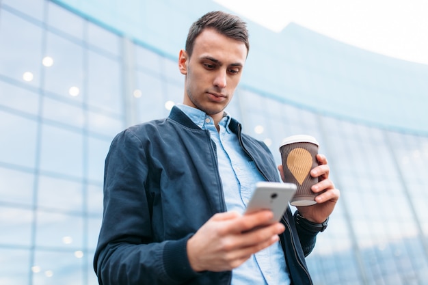 Un hombre con una taza de café de papel, recorre la ciudad, un chico guapo con ropa elegante, con el teléfono en la mano.