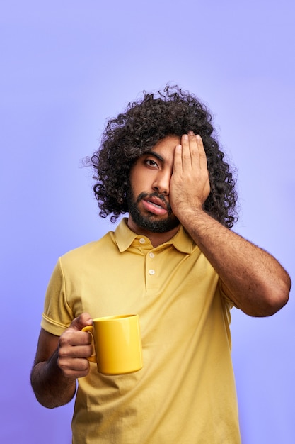 Hombre con una taza de té, el hombre árabe rizado necesita dormir, cerrando un ojo posando en la cámara aislada sobre el espacio púrpura