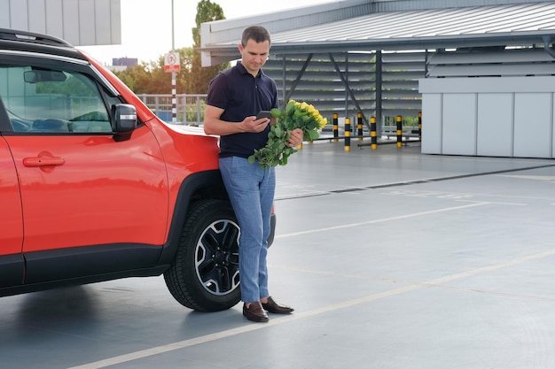 Un hombre con un teléfono en las manos y un ramo de flores en el estacionamiento del aeropuerto