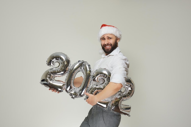 Un hombre tenso con la cabeza en alto con un gorro de Papá Noel de terciopelo rojo se lleva globos plateados con la forma del 2022. Un tipo con barba en una fiesta de Año Nuevo.