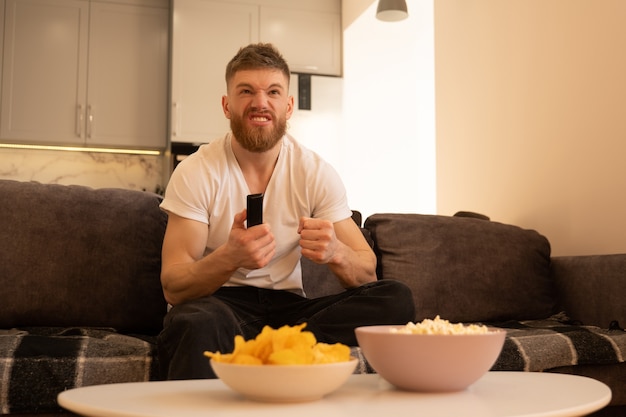 El hombre tenso se sienta en el sofá y ve la televisión o una película. Chico barbudo europeo joven con mando a distancia. Cuencos con patatas fritas y palomitas de maíz en la mesa. Concepto de descanso en casa. Interior del apartamento estudio