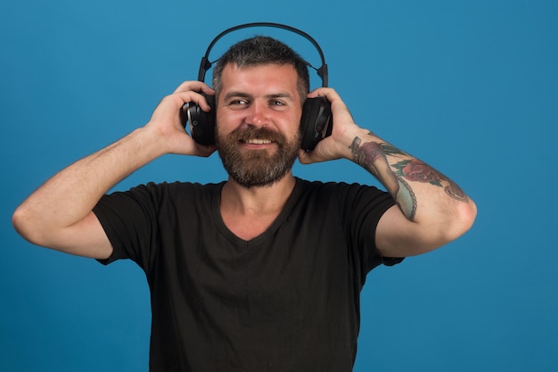 El hombre tiene auriculares con fondo azul. Dj con barba usa auriculares. Placer, música y concepto de estilo de vida creativo. Cantante con barba y cara feliz escucha música.