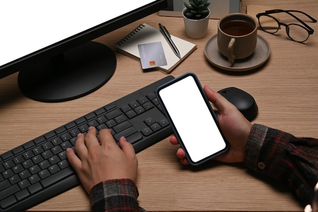 Hombre de tiro recortado sosteniendo un teléfono inteligente y trabajando con la computadora en la oficina en casa