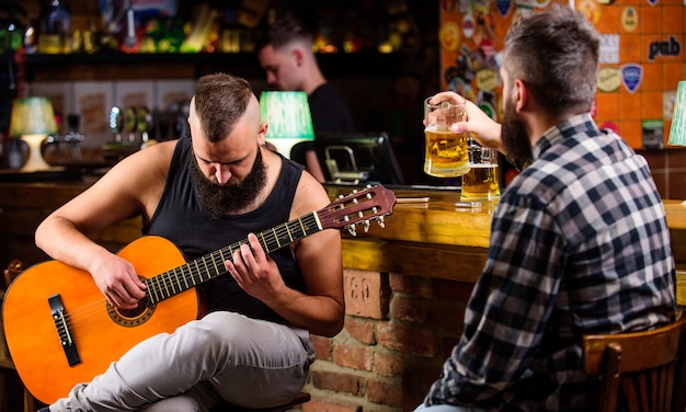 El hombre toca la guitarra en el bar. Viernes relajación en bar. Amigos relajándose en bar o pub. Hipster brutal barbudo pasar ocio con un amigo en el bar. Ocio de hombres de verdad. Amigos alegres se relajan con música de guitarra.