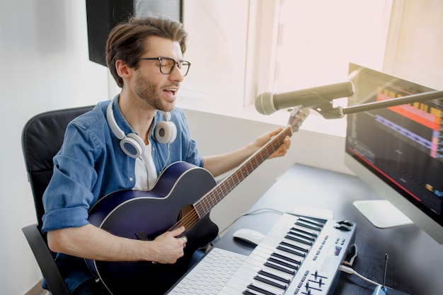 El hombre toca la guitarra y canta y produce una banda sonora electrónica o una pista en un proyecto en casa. Arreglista masculino componiendo canciones en piano midi y equipo de audio en estudio de grabación digital.