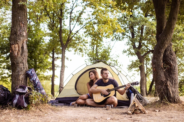 Un hombre toca para una mujer en una guitarra cerca de una carpa al atardecer. Luna de miel en la caminata