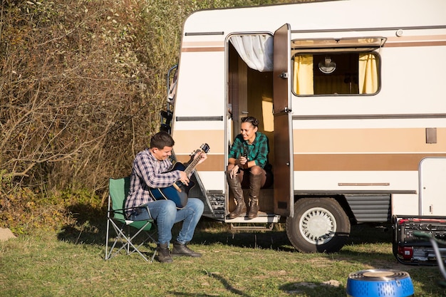 Hombre tocando buena música en la guitarra a su novia frente a su autocaravana retro.